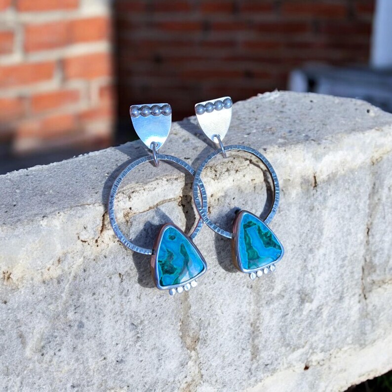 May include: A pair of silver hoop earrings with turquoise and green triangular stones. The earrings have a hammered texture on the hoops and decorative silver accents. The earrings are displayed on a gray stone surface.