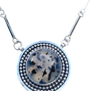 May include: A silver necklace featuring a large, round pendant with a speckled, translucent stone center. The pendant is encircled by a beaded border and a smooth silver frame. The chain has a delicate design.