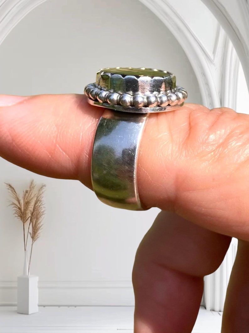 Puede incluir: Un anillo de plata con una parte superior redonda de color dorado. La parte superior tiene un borde de cuentas y una superficie texturizada.
