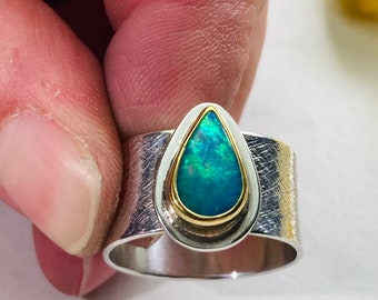 May include: A silver and gold ring with a teardrop-shaped opal gemstone. The ring has a textured band and a smooth, polished finish.