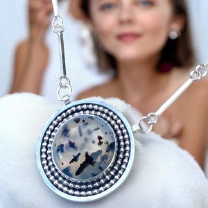May include: A silver pendant necklace featuring a large, round stone with a speckled pattern of black and white. The stone is set in a silver bezel with a beaded border. The necklace has a simple, elegant design.