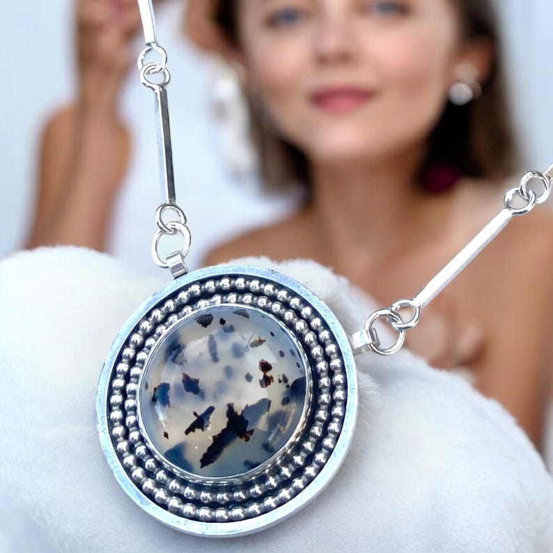 May include: A silver pendant necklace featuring a large, round stone with a speckled pattern of black and white. The stone is set in a silver bezel with a beaded border. The necklace has a simple, elegant design.