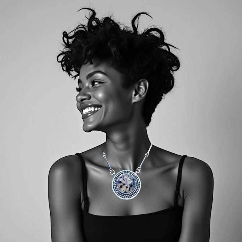 May include: A black and white portrait of a smiling person wearing a silver necklace. The necklace features a large, round pendant with a patterned design in shades of blue, white, and gray. The person is wearing a black tank top.