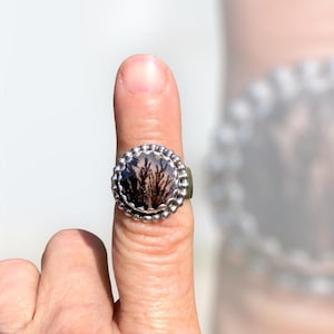Puede incluir: Un anillo de plata con un engaste redondo de bisel con cuentas. El bisel contiene una piedra transparente con un patr&oacute;n de &aacute;gata dendr&iacute;tica en tonos marrones y negros.