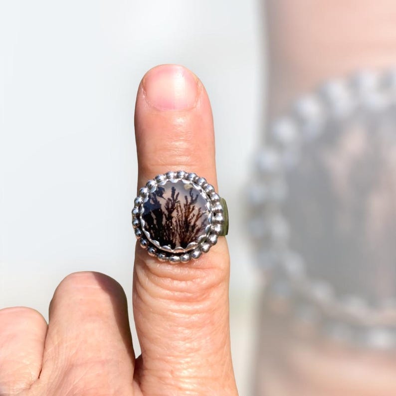 Puede incluir: Un anillo de plata con un engaste redondo de bisel con cuentas. El bisel contiene una piedra transparente con un patr&oacute;n de &aacute;gata dendr&iacute;tica en tonos marrones y negros.