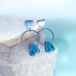 May include: A pair of silver hoop earrings with turquoise-colored triangular stones. The earrings have a textured design on the hoops and small silver details at the top and bottom of the turquoise stones.