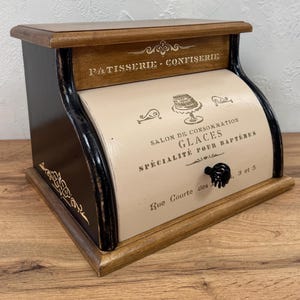 Könnte beinhalten: Ein Brotkasten aus Holz mit Rolltür und einer cremefarbenen Frontplatte mit schwarzer Aufschrift PATISSERIE-CONFISERIE, SALON DE CONSOMMATION, GLACES, SPÉCIALITÉ POUR BAPTÊMES und Rue Courte des, sowie einem kleinen schwarzen Knaufgriff.