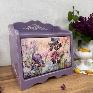 May include: A lavender wooden bread box with a floral iris design on the front. The box has a scalloped top and decorative accents. A silver handle is on the drawer. The background includes a vase of purple flowers and a white dish with cookies.