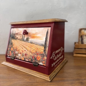 May include: A burgundy wooden bread bin with a hinged door and a decorative landscape scene featuring a field of poppies and a Tuscan villa. The bin has a wooden base and a small red knob. The text "ATELIERS Du Chocolat" is visible.