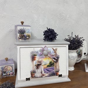May include: A decorative white wooden box with a lavender field scene, featuring a woman in a hat. The box has a gray top and is accompanied by matching lavender-themed containers and a vase with lavender sprigs. The overall theme is rustic and floral.