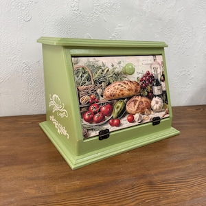 Può includere: Un portapane in legno verde con anta a cerniera. L'anta presenta un'illustrazione colorata di pane, pomodori, aglio e vino. Il portapane ha un motivo floreale bianco decorativo e poggia su una superficie in legno.