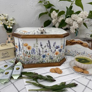Può includere: Una scatola per il pane bianca con base e coperchio in legno, decorata con un motivo floreale con fiori gialli, blu e viola. Un piccolo pomello in ceramica è sopra il coperchio. Altri oggetti includono una burriera, biscotti e un portapane.