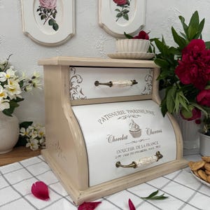 May include: A cream-coloured wooden bread bin with two drawers and a curved front. The front of the bin has the text "PATISSERIE PARISIENNE Chocolat Churros". Two framed rose prints hang above the bin.