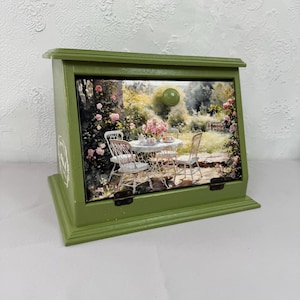 Può includere: Una scatola di legno verde con coperchio incernierato e una scena paesaggistica decorativa sul davanti. La scena raffigura un giardino con un tavolo e sedie. La scatola ha un pomello verde e cerniere in metallo.