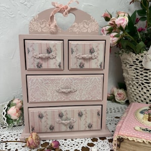 Könnte beinhalten: Eine kleine, rosa Holzkommode mit vier Schubladen. Die Schubladen weisen Blumen- und Streifenmuster mit verzierten Griffen auf. Ein herzförmiger Ausschnitt und ein rosa Band zieren die Oberseite. Die Kommode ist mit einem floralen Design verziert.