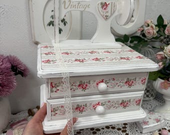 May include: White vintage-style sewing machine-shaped jewelry box with two drawers. The box is decorated with floral patterns in red and pink. The word "Vintage" is printed on the machine's arm. A white lace ribbon is draped over the box.