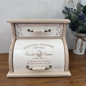 Puede incluir: Una panera de madera de dos niveles, de color blanco roto, con un acabado envejecido. El cajón superior tiene un diseño decorativo y un tirador de cerámica. La sección inferior presenta el texto "Patisserie Parisienne" y una ilustración de magdalena. Las asas son de metal.