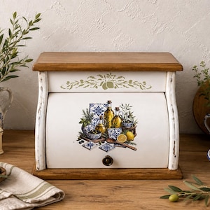 Könnte beinhalten: Ein Brotkasten aus Holz mit Rolltür, weißer Oberfläche sowie Ober- und Unterseite aus Naturholz. Die Vorderseite zeigt eine farbenfrohe Illustration von Olivenölflaschen, Zitronen und blau-weißen Keramikfliesen mit dekorativen Mustern.