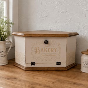 May include: A hexagonal wooden bread box with a cream-colored front panel featuring the text "BAKERY PIES CAKES COOKIES 1913" in brown lettering. The box has a dark wood top and base, a single black knob, and two black metal hinges at the bottom.