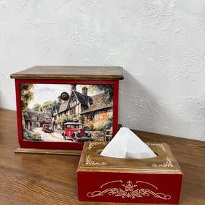 May include: A red and brown wooden tissue box and matching box with a hinged lid. The boxes feature a vintage village scene with cars. The tissue box has a decorative gold design on the front.