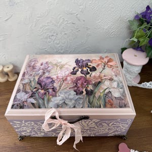 May include: A rectangular wooden box with a floral iris design on the lid. The box features a light purple base with a decorative pattern and a pink ribbon tied in a bow. The box has dark metal hardware and small feet.