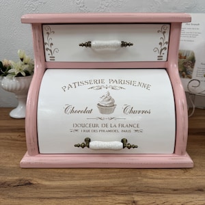 May include: A pink and white wooden bread box with a top drawer and a roll-top door. The door features the text PATISSERIE PARISIENNE, Chocolat, Churros, and DOUCEUR DE LA FRANCE 1 RUE DES PYRAMIDES, PARIS, with a cupcake graphic and metal handles.