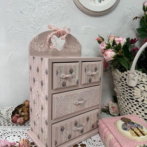 Può includere: Un portagioie in legno rosa con quattro cassetti, decorato con motivi floreali e a righe. La parte superiore presenta un ritaglio a forma di cuore e un fiocco decorativo. La scatola è appoggiata su un tavolo con una tovaglia di pizzo, circondata da rose e un libro decorativo.