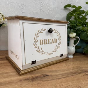May include: A rectangular wooden bread box with a white painted finish and a natural wood-toned top and base. The front door features a dark metal knob, a beige laurel wreath graphic, the text BREAD, and the year 1820 printed in beige.
