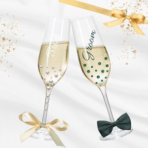 May include: Two champagne flutes clinking together, one with "Bride" etched on it and adorned with pearls, the other with "Groom" and green polka dots. A dark green bow tie and a gold ribbon with a bow add to the celebratory theme.