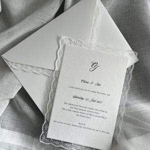 Può includere: Un invito di nozze bianco con bordo in pizzo e busta abbinata. L'invito presenta una scrittura elegante e i nomi "Olivia & Jan", insieme alla data "Samstag, 23. Juli 2027".