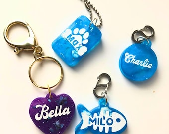 May include: Four personalized pet ID tags with different shapes and colors. A blue rectangular tag with a paw print and the name "Max", a blue round tag with the name "Charlie", a purple heart-shaped tag with the name "Bella", and a blue fish-shaped tag with the name "Milo".