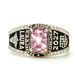 May include: A gold class ring featuring a central cushion-cut pink gemstone surrounded by black enamel. The band is engraved with "LAURA", "NOUVEAU HIGH SCHOOL", and "2027", accented by a caduceus symbol, a crest, and rows of small clear stones.