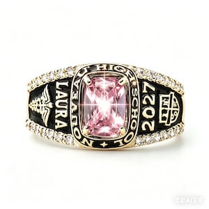 May include: A gold-toned class ring with a large, rectangular pink gemstone at its center. The ring features the name "LAURA" and the year "2027" in white lettering on a black background, surrounded by small, clear gemstones. The words "NOUVEAU HIGH SCHOOL" are also visible.