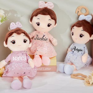 May include: Three plush dolls with brown hair and bows, wearing dresses. The dolls are in pink and blue, with the names "Nancy", "Jackie", and "Angie" printed on their dresses. The dolls are sitting on a white surface.