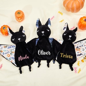 Puede incluir: Tres mantas de bebé con forma de murciélago de peluche negro con nombres personalizados bordados en rosa y dorado. Las alas presentan un estampado temático de Halloween. Las calabazas y los caramelos están esparcidos.