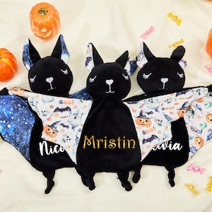 Puede incluir: Tres juguetes de murciélago de peluche negro con nombres personalizados, cada uno con un diseño de ala único. Un ala tiene un patrón de galaxia, y las otras tienen un estampado de Halloween con calabazas y fantasmas. Los murciélagos están rodeados de calabazas y caramelos.