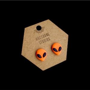 May include: A pair of orange alien face stud earrings with black eyes on a brown hexagonal card with the text "Nocturne Studios".