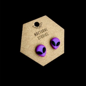 May include: A pair of purple alien head stud earrings with black eyes. The earrings are displayed on a brown card with the text "Nocturne Studios".