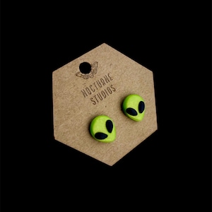 May include: A pair of green alien head stud earrings with black eyes. The earrings are displayed on a hexagonal card with the text "Nocturne Studios".