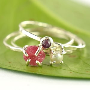 Pode incluir: Três anéis de prata com pedras preciosas. O primeiro anel tem um rubi rosa, o segundo anel tem uma pequena pedra preciosa roxa e o terceiro anel tem uma pérola branca.