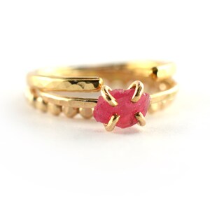 Op de afbeelding: Een gouden ring met een roze edelsteen in het midden. De ring is gemaakt van twee banden, een met een gehamerde textuur en de andere met kleine kralen.
