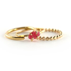 Op de afbeelding: Twee gouden ringen, een met een getextureerde band en de andere met een kralenband, zijn op elkaar gestapeld. Een kleine, ruwe roze edelsteen is in de getextureerde band gezet.