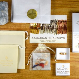 Puede incluir: Una imagen de plano de una variedad de art&iacute;culos de joyer&iacute;a, incluyendo collares, pulseras y anillos, empaquetados en cajas blancas y peque&ntilde;as bolsas. La joyer&iacute;a est&aacute; hecha por Aquarian Thoughts, una empresa que se especializa en joyer&iacute;a artesanal. La imagen tambi&eacute;n incluye una peque&ntilde;a pieza de cristal de selenita azul.