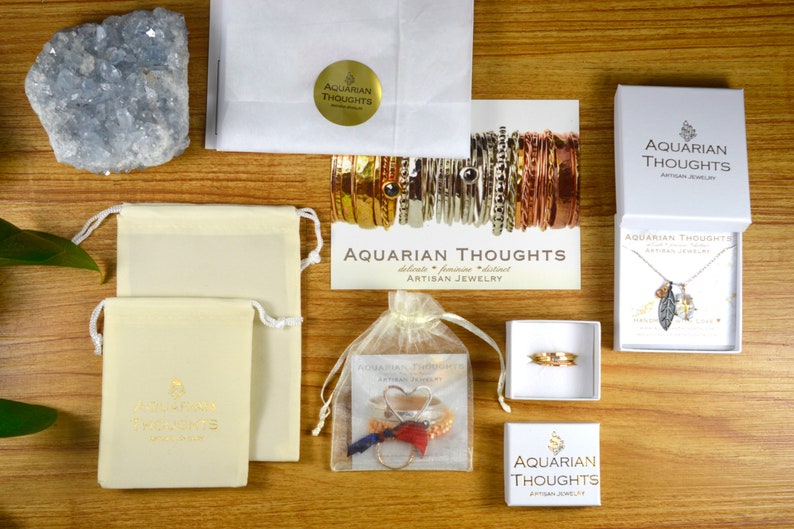Puede incluir: Una imagen de plano de una variedad de art&iacute;culos de joyer&iacute;a, incluyendo collares, pulseras y anillos, empaquetados en cajas blancas y peque&ntilde;as bolsas. La joyer&iacute;a est&aacute; hecha por Aquarian Thoughts, una empresa que se especializa en joyer&iacute;a artesanal. La imagen tambi&eacute;n incluye una peque&ntilde;a pieza de cristal de selenita azul.