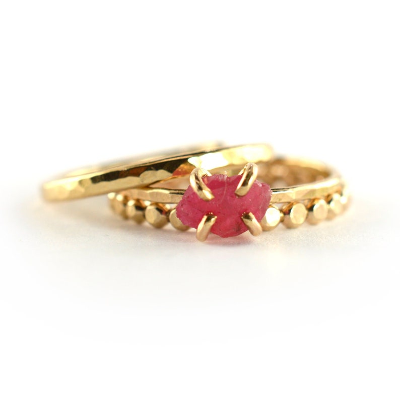 Op de afbeelding: Een set van drie gouden ringen met een roze edelsteen. De ringen zijn op elkaar gestapeld. De bovenste ring heeft een gehamerde afwerking, de middelste ring heeft een kralenontwerp en de onderste ring is een eenvoudige band.