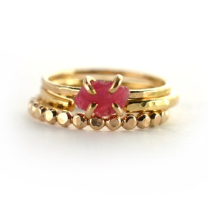 Op de afbeelding: Drie gouden ringen gestapeld op elkaar. De bovenste ring heeft een roze edelsteen. De onderste ring is een kralenring.
