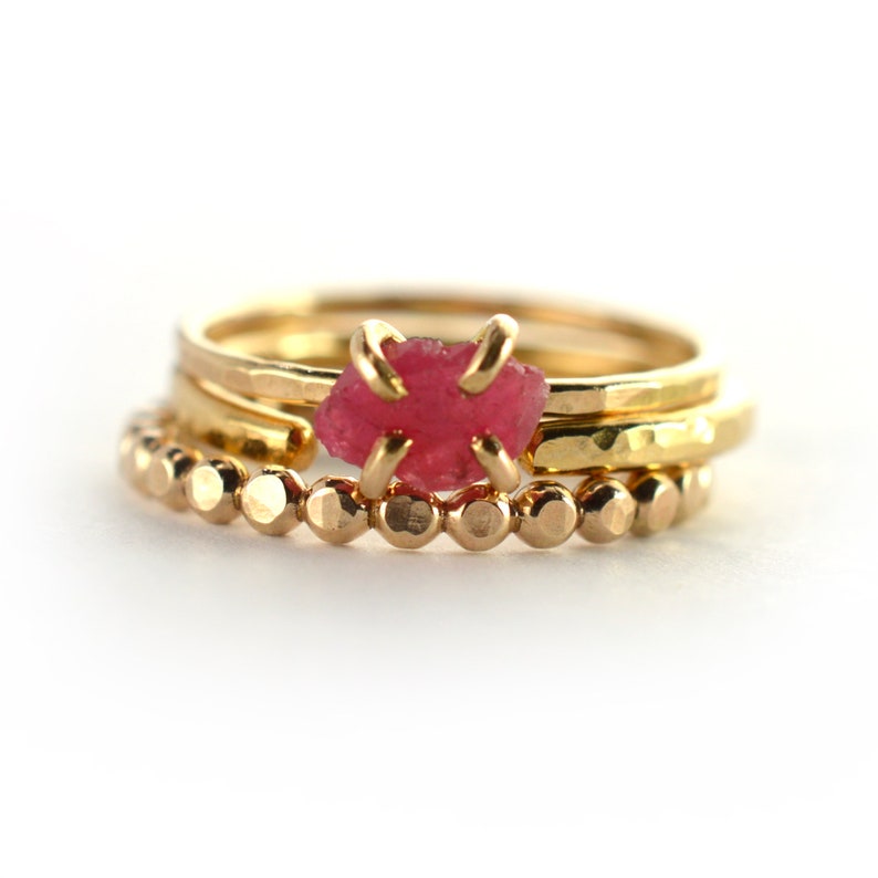 Op de afbeelding: Drie gouden ringen gestapeld op elkaar. De bovenste ring heeft een roze edelsteen. De onderste ring is een kralenring.