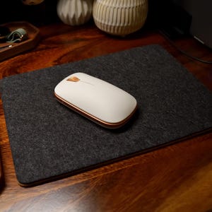May include: A white wireless computer mouse with gold trim rests on a dark gray felt mousepad. A wooden keyboard and tray with accessories are also visible on a wooden desk. The setup suggests a workspace or home office.