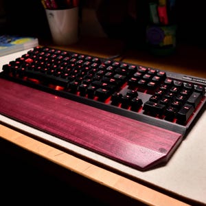 May include: A black mechanical keyboard with red backlit keys and a wooden wrist rest. The keyboard sits on a light-colored desk mat, with a black mouse to the right. The wrist rest is a deep reddish-purple color.