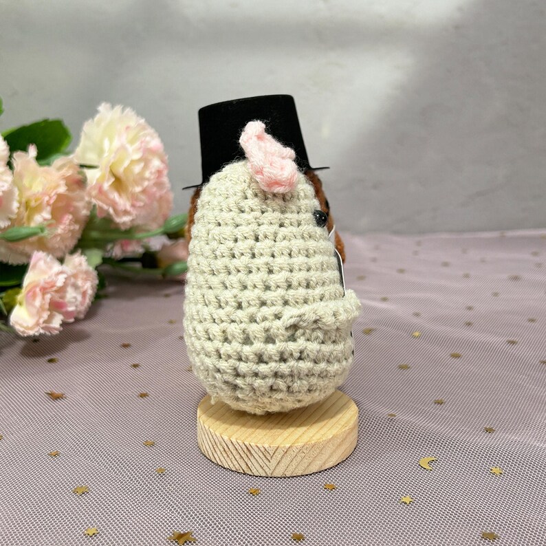May include: A crocheted figure of a hedgehog wearing a black top hat and a light pink ear. The hedgehog is made of beige yarn and sits on a small wooden base. The background includes pink flowers.